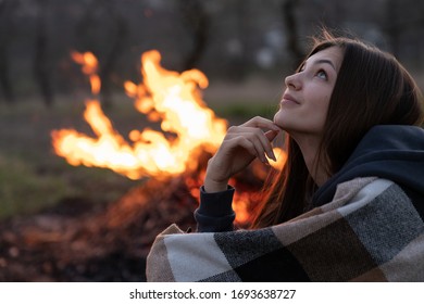 Dreamy romantic brunette girl in plaid looking up, big campfire on the background, spring time