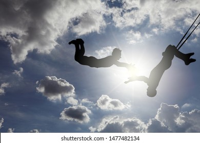 Dreamy Silhouette Of A Pair Of Trapeze Artists Swinging Through A Blue Cloudy Sky. 