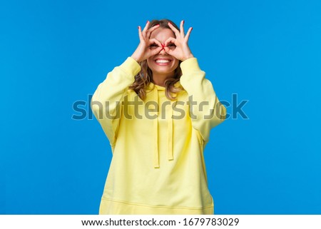 Dreamy and silly girl looking through fingers with happy playful expression, make okay gestures form glasses with hands and smiling carefree, look far away, searching something, blue background