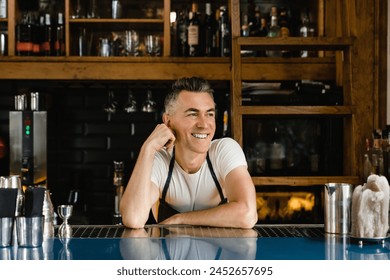 Dreamy smiling caucasian mature bartender small business owner barista waiter leaning on the bar counter in blue apron in restaurant - Powered by Shutterstock