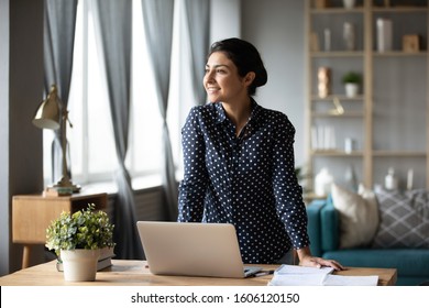 Dreamy Smiling Young Indian Woman Student Online Teacher Freelancer Looking Away Dreaming About Professional Future Success Hope For New Opportunity Concept At Home Office Stand At Table With Laptop