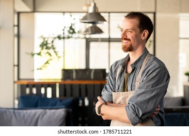 Dreamy successful young bearded coffee shop owner in apron standing with crossed arms and looking into distance - Shutterstock ID 1785711221