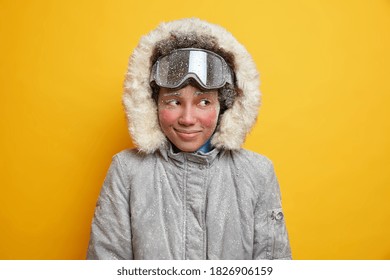 Dreamy Winter Woman With Red Frozen Face Enjoys Holiday Mountain Resort During Cold Good Snow Day Covered By Snowflakes Dressed In Warm Jacket With Hood Wears Ski Goggles Likes Extreme Sport.