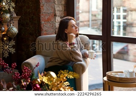 Dreamy woman in sweater sits and looks in armchair by window in loft style. Girl greets morning of Christmas with mug of hot chocolate or cocoa with marshmallows in living room decorated for holiday