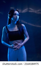 Dreamy Woman Wearing Black Headphones Standing, Squeezing Hands Inside. Front View Of Tranquil Female Dj In Crop Top Listening To Music, Lighted By Dark Blue Light In Studio. Concept Of Gesturing. 