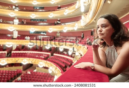 Dreamy young adult woman sitting on balcony in theater hall during intermission, examining interior in anticipation of continuation of theatrical production..
