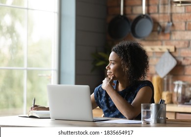 Dreamy young African American woman distracted from laptop work look in window distance dreaming thinking. Happy 20s biracial female lost in thoughts planning or visualizing at home office.