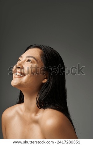 dreamy and young asian woman with brunette hair and acne prone skin smiling on grey background