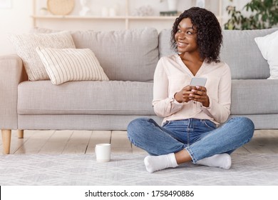 Dreamy Young Black Girl Relaxing On Floor At Home With Smartphone And Coffee, Enjoying Weekend, Copy Space