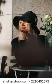 Dreamy Young Blond Woman In A Floppy Hat And A Long Sleeved Balck Dress, Sitting Behind Cafe Table Outside, Looking Afar, Smiling.