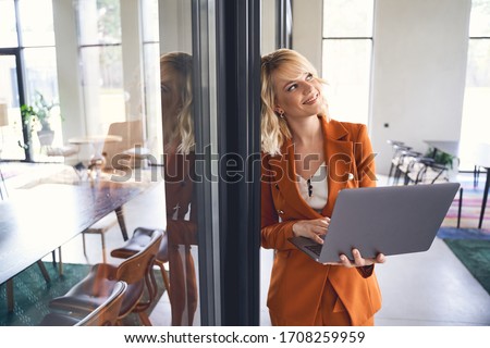 Dreamy young business lady in a stylish jacket leaning against a wall in the office
