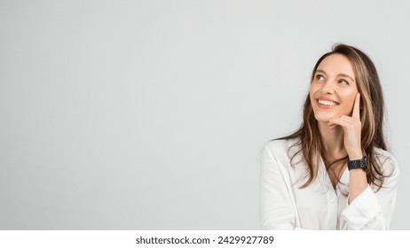 Dreamy young european woman with a joyful smile looking upwards, resting her face on her hand, wearing a white blouse, jeans, and a smartwatch, against a spacious grey background - Powered by Shutterstock