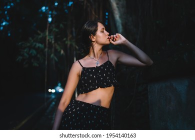 Dreamy young female with dark hair in trendy outfit standing at dusk near forest road in late evening and looking away