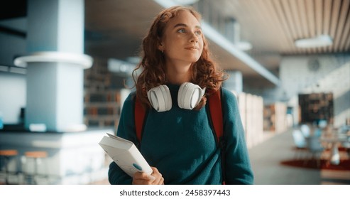 Dreamy Young Female Student Wandering Through Modern Library With Books and Headphones, Wearing Casual Green Sweater and Carrying Red Backpack, Looking Upward With Curious Expression - Powered by Shutterstock