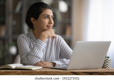 Dreamy young Indian woman sit at desk at home office work on laptop thinking or pondering. Pensive millennial mixed race female distracted from computer job look in distance visualizing planning.
