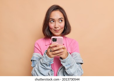 Dreamy young pretty Asian woman with dark hair holds mobile phone has thoughtful expression thinks about received message wears pink t shirt denim jacket looks away isolated over beige background.