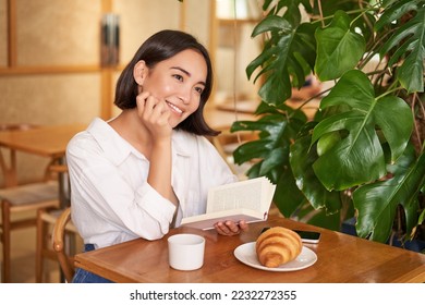 Dreamy young smiling asian woman reading book, sitting in cafe, eating croissant and drinking coffee in cozy interior. - Powered by Shutterstock