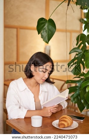 Dreamy young smiling asian woman reading book, sitting in cafe, eating croissant and drinking coffee in cozy interior.