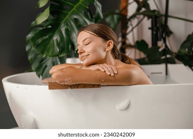 Dreamy young woman leaning on bathtub side and resting in the bathroom decorated with tropical plant. Wellness, beauty and care concept - Powered by Shutterstock