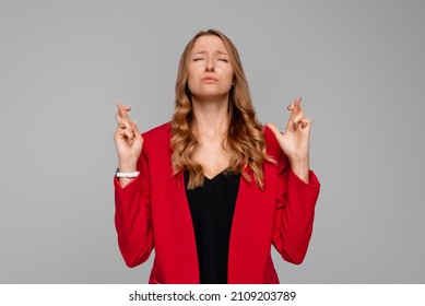 Dreamy Young Woman Makes Wish With Crossed Fingers, Close Eyes And Pray, Begging God For Dream Come True, Yearning To Achieve Goal, Standing In Red Blazer Against Gray Background