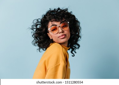 Dreamy young woman in sunglasses looking at camera. Front view of interested curly girl isolated on blue background. - Powered by Shutterstock