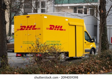 DRESDEN, GERMANY - 27. November 2021: Yellow DHL Delivery Car Parking In Front Of Buildings. Tree And Bushes Are In The Foreground. Delivering Parcels And Packages To Customers At Home.