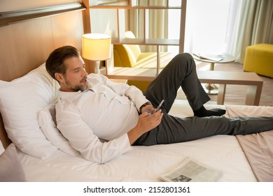 Dressed bearded man lying on a hotel bed using his smartphone