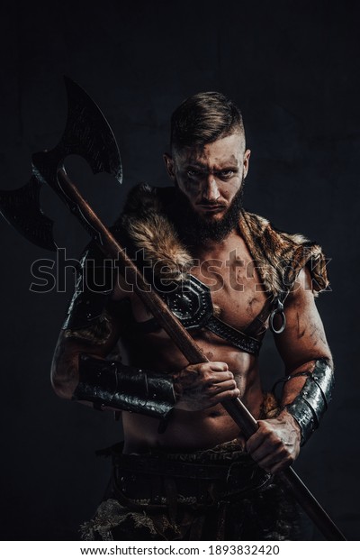 Dressed in dark armour with fur dirty and\
awesome viking fighter with two handed axe on his shoulder poses in\
dark background.