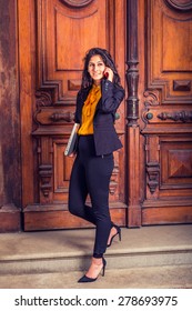 Dressing In Black Blazer, Orange Shirt, Pants, Heels, A Young East Indian American Business Woman Walking Down Vintage Style Office Door Way, Carrying Laptop Computer, Making Call On Her Mobile Phone.