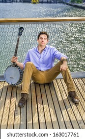 Dressing In A Light Blue Shirt, Dark Yellow Jeans And Brown Boot Shoes, Holding A Banjo, A Young Musician Is Sitting On The Deck Against A Fence, Relaxing /Musician Relaxing Outside