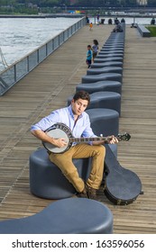 Dressing In A Light Blue Shirt, Dark Yellow Jeans And Brown Boot Shoes, A Young Musician With An Instrument Box Is Sitting On A Modern Style Bench, Playing A Banjo. / Play Music Outside