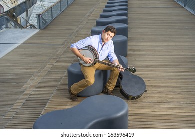 Dressing In A Light Blue Shirt, Dark Yellow Jeans And Brown Boot Shoes, A Young Musician With An Instrument Box Is Sitting On A Modern Style Bench, Playing A Banjo. / Play Music Outside 