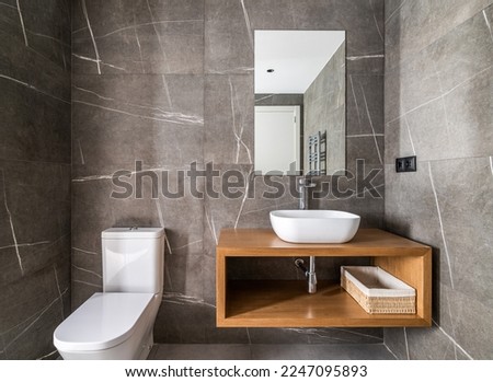 Dressing room with walls made of dark gray marble with white touches, small ceramic oval sink on wooden cabinet and mirror on wall that reflects radiator and front door to room. Stylish bathroom.