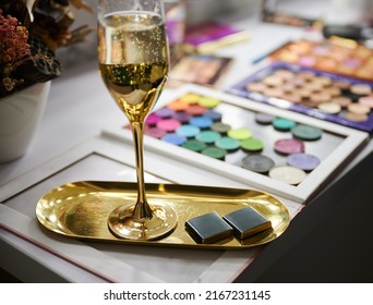 Dressing table with makeup eyeshadow palette, glass of alcoholic drink and corporate business chocolate on gold tray. Champagne, sweets and makeup tools in visage studio.