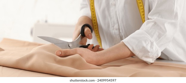 Dressmaker cutting fabric with scissors at table in workshop, closeup. Banner design