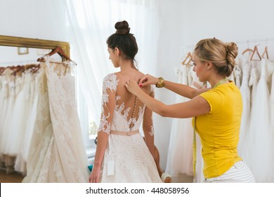 Dressmaker Helping The Bride To Put Her Wedding Dress On In Clothes Shop.