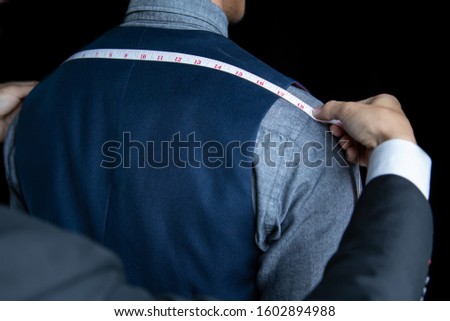 The dressmaker was measuring the width of the back of the man wearing a blue suit on black background.