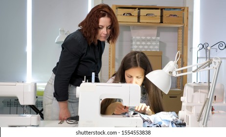 Dressmaker Teaches New Employee To Work In Workshop. Cutting And Sewing Courses. Seamstress Teacher Controls Looks How Student Sew On Sewing Machine In Workshop. Traineeship Concept. Probation Period.