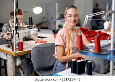 Dressmakers women sewing clothes on sewing machine in factory together, at work. Attractive ladies enjoy tailoring, making clothes, blonde caucasian craftswoman looking at camera smiling - Powered by Shutterstock