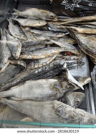 Dried fish are piled tightly inside market display case, showing silvery scales, rough skin, and pale brown tones. Fresh produce peeks through gaps, suggesting food retail and seafood trade