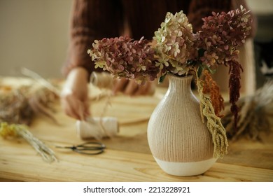 Dried hydrangea flowers in vase on background of woman arranging dried grass in wreath on wooden table. Making stylish autumn wreath on rustic table. Fall decor and arrangement in farmhouse - Shutterstock ID 2213228921