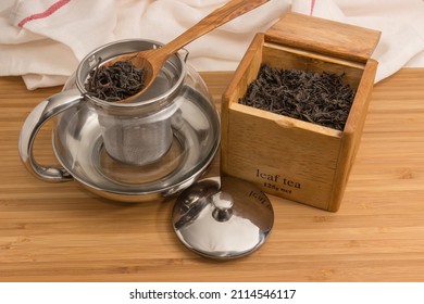 Dried Large Leaves Of Black Tea In The Wooden Spoon On An Empty Glass Teapot With Removed Lid, Same Black Tea In An Open Wooden Box 
