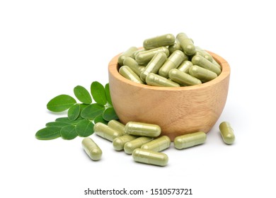 Dried Moringa leaves powder capsules in wooden bowl with green leaves isolated on white background.