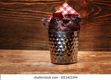 Dried Peppered Beef Jerky Cut In Strips In A Metal Bucket On Wooden Background. Serving The Dish In A Restaurant