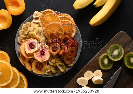 Dried sliced plums, kiwi, peach fruits chips in plate shape of heart on black background, home drying. Snack vegan free sugar food. View from above.