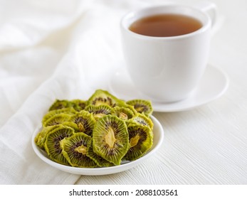 Dried Thin Slices, Kiwi Fruit Chips On White Plate And Cup Of Tea. Healthy Food And Snacks. Fruits Are Bright Orange In Color. Side View Close Up, Light Background. Minimalism