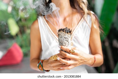 Dried white sage smoke, speckled stick burning in soft focus with bokeh, blurring aroma close-up. Girl's hands hold dried leaves