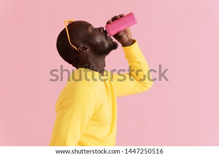Drink. Black man drinking soft drink on pink background. Colorful portrait of happy smiling african american male model in yellow fashion clothes enjoying soda from pink can in studio 