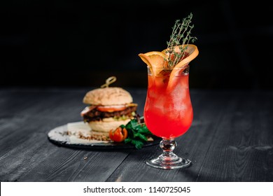 Drink And Food. Red Citrus Cocktail With Orange Grapefruit And Ice With Burger On The Black Wood Table