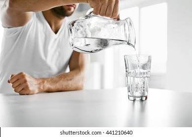 Drink Water. Close Up Of Handsome Young Man Pouring Fresh Pure Water From Pitcher Into A Glass In Morning In Kitchen. Beautiful Athletic Male Model Feeling Thirsty. Healthy Nutrition And Hydration